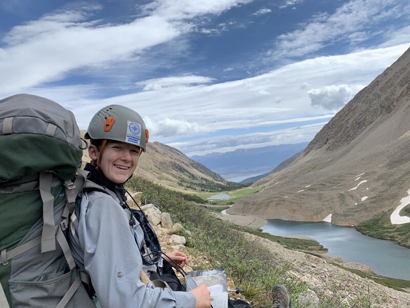A person wearing a helmet and a large backpack is sitting on a rocky hillside, smiling at the camera. In the background, there's a scenic mountain valley with a lake and patches of snow. The sky is partly cloudy, suggesting a bright day for hiking or climbing. The overall impression is one of adventure and enjoying the outdoors.